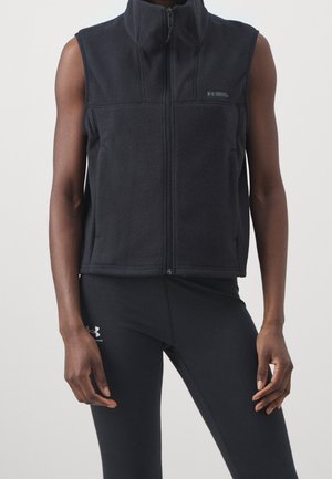 Osoba ubrana w czarną kamizelkę polarową bez rękawów z zamkiem błyskawicznym oraz czarne legginsy Under Armour na jednolitym tle.