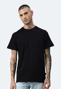 Schwarzes Baumwoll-T-Shirt mit kurzen Ärmeln und klassischem Rundhalsausschnitt. Das Modell zeigt tätowierte Arme vor einem hellen Hintergrund.
