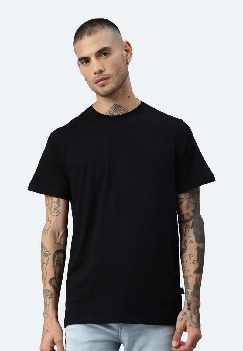 Schwarzes Baumwoll-T-Shirt mit kurzen Ärmeln und klassischem Rundhalsausschnitt. Das Modell zeigt tätowierte Arme vor einem hellen Hintergrund.