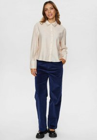 Gai beige krūšu pogu krekls ar apkakli, garām piedurknēm; saskaņots ar tumši zilām korduroja biksēm; modelis stāv ar vienu roku kabatā.