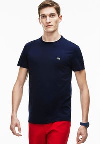 Temno modra bombažna majica s krožnim izrezom, kratkimi rokavi in majhnim zelenim logotipom Lacoste na prsih. Nosi se z rdečimi hlačami.