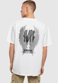 Hvid bomulds t-shirt med et stort, detaljeret grafisk motiv af sorte og grå englevinger, hvor ordet "HOPE" er fremtrædende.
