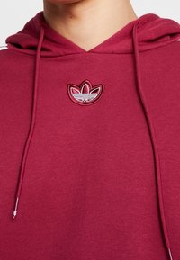 Bordeauxfärgad hoodie med justerbar snörning i huvan. Har en broderad logotyp med tre blad på bröstet. Mjuk, texturerad tyg.