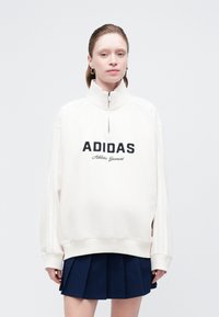 Felpa bianca con zip e la scritta "ADIDAS" in grassetto nero. Caratterizzata da un colletto alto, spalle scese e orlo a coste.
