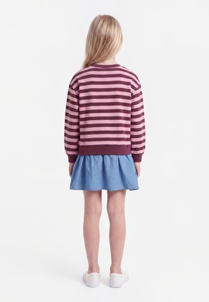 Barn med langt blondt hår iført en pink og mørkerød stribet sweater, blå nederdel og hvide sko, der står med ryggen mod beskueren.