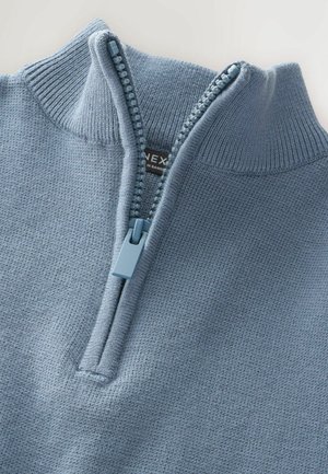 Gilet zippé bleu clair avec un col haut, un col à côtes et une fermeture éclair bleue contrastante. Texture tricotée lisse avec des détails subtils.