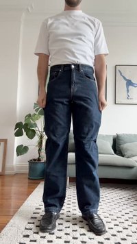 Jeans de ganga azul escuro com um corte reto, quatro bolsos e fecho com botão. Combinados com uma t-shirt branca lisa e sapatos pretos.