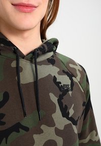 Hnědá a zelená maskáčová mikina s kapucí na šňůrku. Disponuje černými akcenty a malým logem Nike na hrudi. Příjemný na dotek.