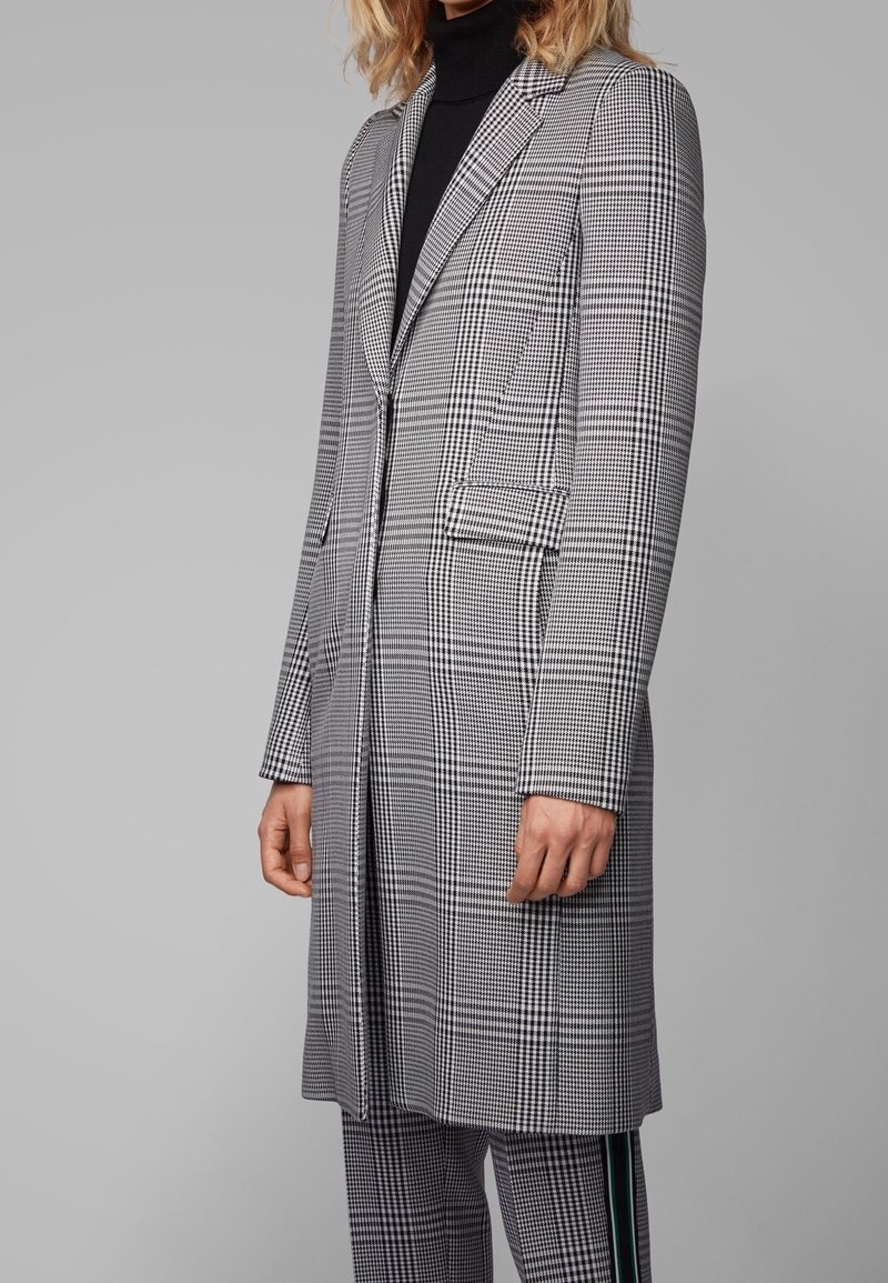 Abrigo a cuadros en gris y negro con una silueta larga, cuello de muesca, bolsillos laterales y una textura suave. Llevado sobre un suéter de cuello alto negro.