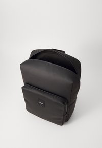 Zaino nero in materiale sintetico liscio, di forma strutturata, con manico superiore, tasca anteriore con zip e discreto dettaglio del logo sul davanti.