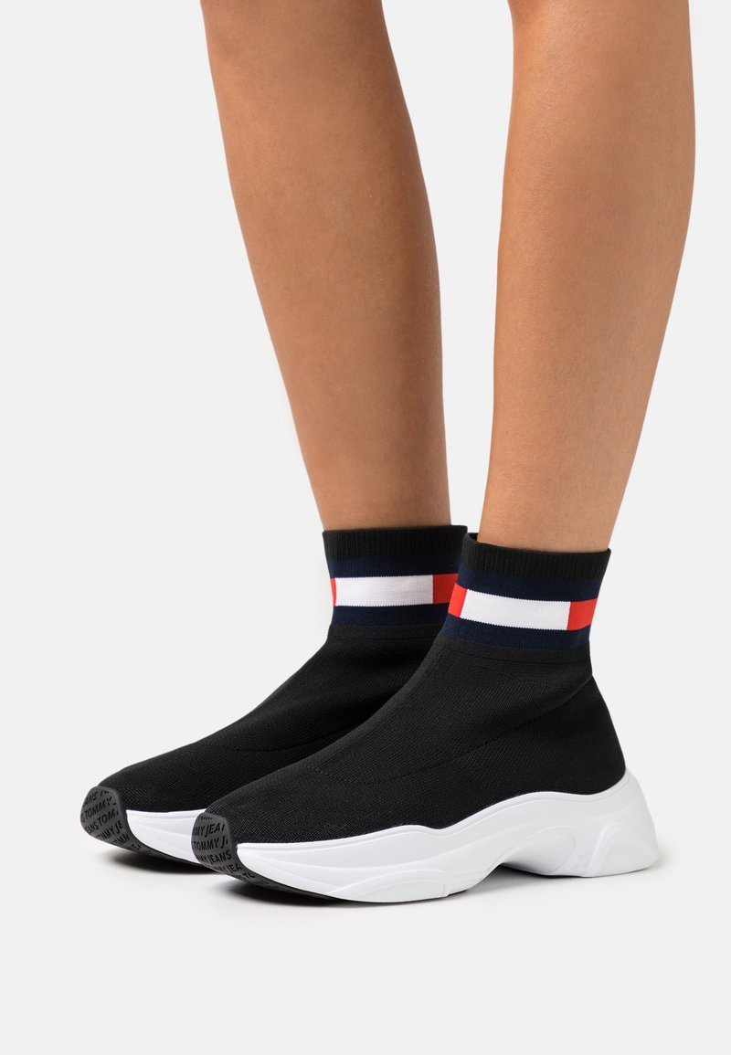 Baskets style chaussettes noires avec semelles épaisses blanches et bandes rouges, blanches et bleues autour de la cheville sur des jambes à peau claire sur fond blanc.