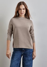 Nainen, jolla on suorat ruskeat hiukset, pitää yllään vaaleanbeigeä pitkähihaista paitaa ja sinisiä farkkuja, seisten yksinkertaista vaaleanharmaata seinää vasten.