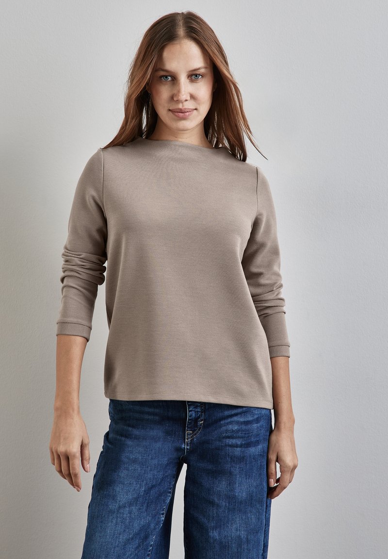 Nainen, jolla on suorat ruskeat hiukset, pitää yllään vaaleanbeigeä pitkähihaista paitaa ja sinisiä farkkuja, seisten yksinkertaista vaaleanharmaata seinää vasten.