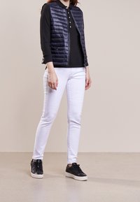 Marinblå vadderad väst över en svart långärmad polo, tillsammans med vita jeans och svarta sneakers. Slät väv, dragkedjestängning och figursydd design.