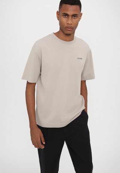 Beige bomull T-skjorte med rund hals, korte ermer og en liten svart "CLOSED"-logo på brystet. Brukt med svarte bukser.