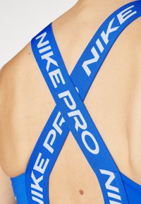 Zils sporta krūšturis ar krusteniskām siksnām. Siksnām ir balts "NIKE PRO" teksts. Gluda auduma, cieši pieguļošs dizains, izceltas plecu detaļas.