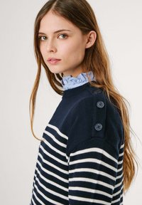 Maglione a righe blu navy e bianche realizzato in tessuto lavorato a maglia, con bottoni decorativi sulla spalla e un colletto arricciato di colore blu chiaro sotto.