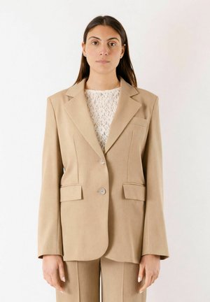 Donna che indossa un blazer beige su misura e pantaloni coordinati sopra una top bianca in pizzo, in piedi contro uno sfondo chiaro e semplice.