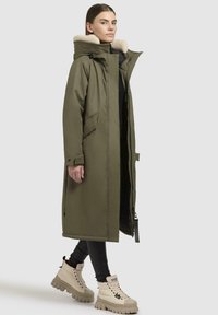 Lange olijfgroene parka met een nepbont kraag, voorzien van een rits aan de voorkant, zijzakken en elastische manchetten, gedragen over zwarte leggings en crèmekleurige laarzen.