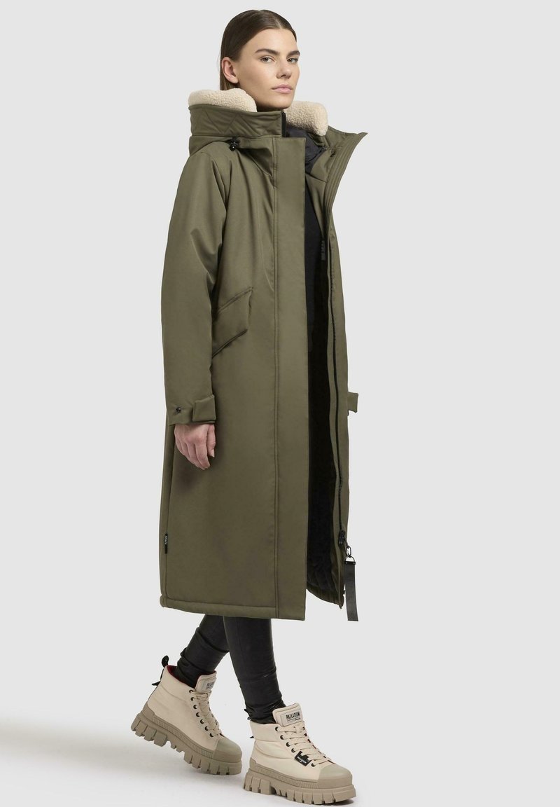 Lange olijfgroene parka met een nepbont kraag, voorzien van een rits aan de voorkant, zijzakken en elastische manchetten, gedragen over zwarte leggings en crèmekleurige laarzen.