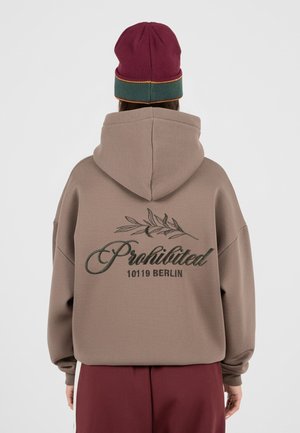 Personne portant un sweat à capuche taupe avec « Prohibited 10119 Berlin » et un motif de branche dans le dos, associé à un bonnet bordeaux et vert.