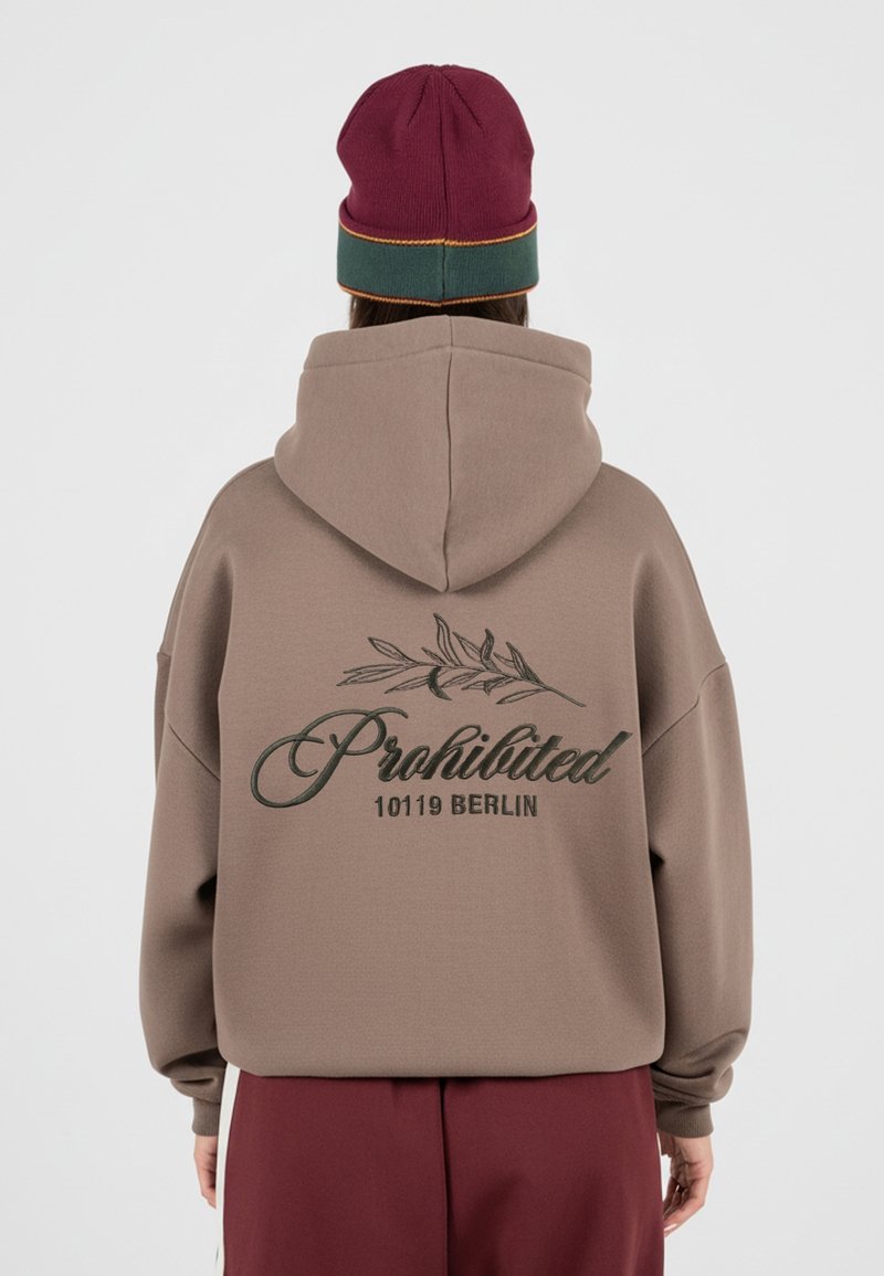 Personne portant un sweat à capuche taupe avec « Prohibited 10119 Berlin » et un motif de branche dans le dos, associé à un bonnet bordeaux et vert.