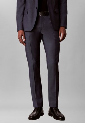 Homme portant un pantalon de costume formel gris foncé, ceinture noire, chaussures noires, debout devant un fond gris clair uni.
