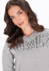 Maglione grigio con scollatura a V, decorato con dettagli in paillettes metalliche in un motivo a onde sulla parte superiore del petto. Texture di maglia morbida.
