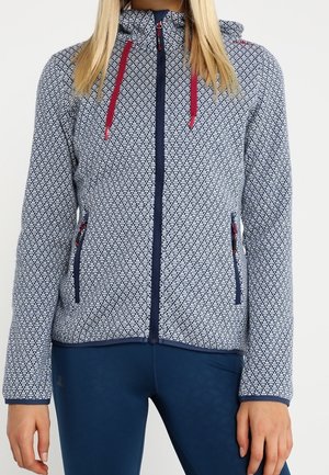 Mujer con sudadera con capucha con cremallera estampada, cordones rojos y mallas azul marino, de pie con los brazos relajados a los lados.
