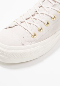 Primo piano di una sneaker in suede beige con bordi scallop, occhielli dorati, lacci bianchi e una suola in gomma bianca testurizzata.
