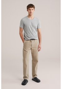 Mladý muž stojaci s rukami v kapsách, oblečený v svetlosivom tričku s výstrihom do V, béžových nohaviciach, čiernom opasku a čiernych slip-on topánkach.