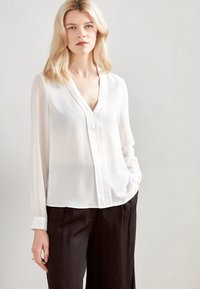Blusa bianca a maniche lunghe con scollo a V profondo e chiusura frontale con bottoni. Realizzata in un tessuto leggero e semi-trasparente. Abbinata a pantaloni scuri.