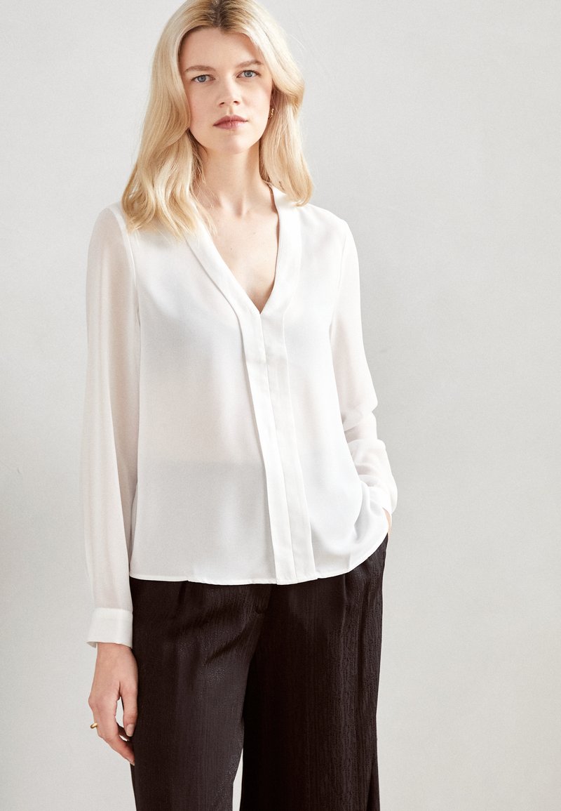 Blusa bianca a maniche lunghe con scollo a V profondo e chiusura frontale con bottoni. Realizzata in un tessuto leggero e semi-trasparente. Abbinata a pantaloni scuri.