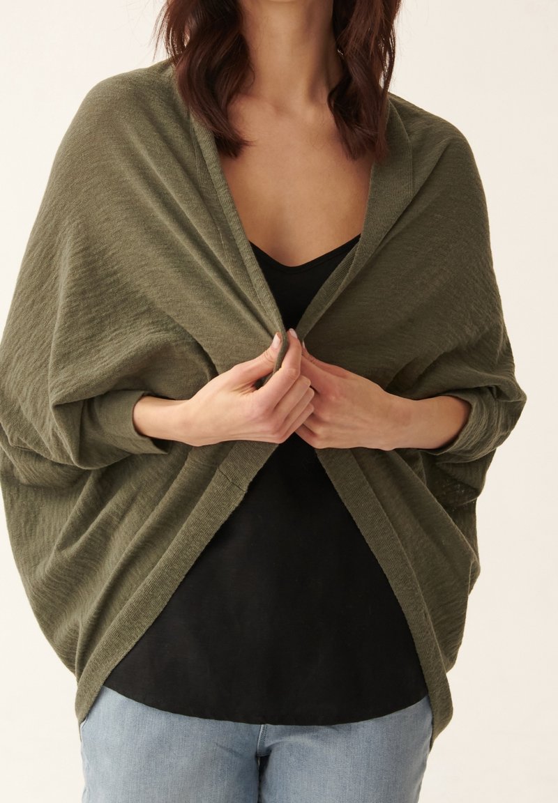 Olijfgroene, lichte wikkel gemaakt van textuurstof, die losjes over een zwart topje valt, met brede mouwen en een open voorkant.