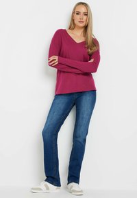 Magenta langmouw V-hals top gecombineerd met blauwe rechte jeans en witte sneakers met beige accenten. Soepele stoftextuur, casual ontwerp.