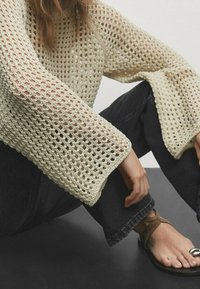 Hellbeige gehäkelter Pullover mit weiten, Glockenärmeln und offener Strickmuster, kombiniert mit dunklen Jeans und flachen Sandalen.
