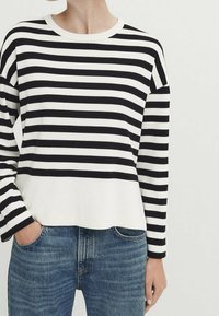 Henkilö, jolla on päällä mustavalkoinen vaakaraitainen pitkähihaispaita, jonka helma on hieman työntötyönsä sinisiin farkkuihin, yksi käsi taskussa.