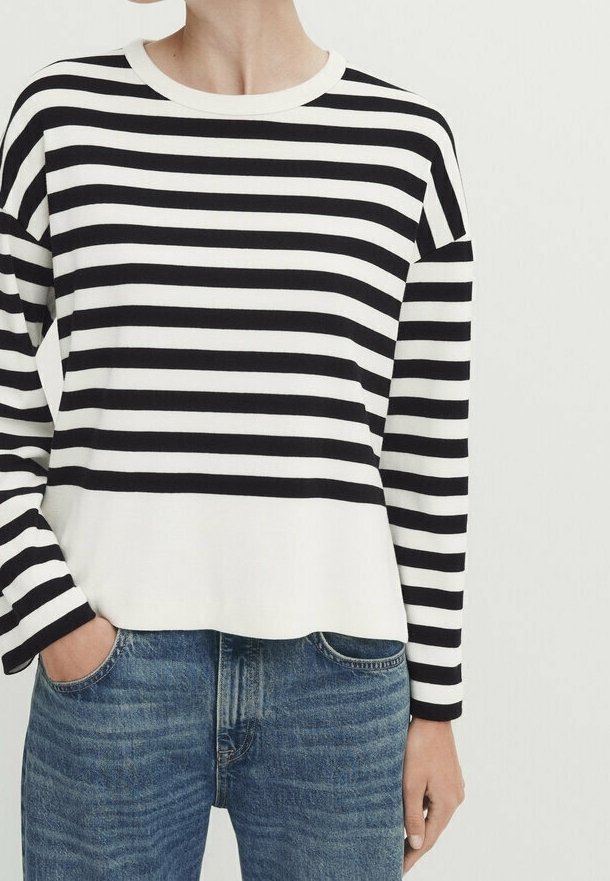 Henkilö, jolla on päällä mustavalkoinen vaakaraitainen pitkähihaispaita, jonka helma on hieman työntötyönsä sinisiin farkkuihin, yksi käsi taskussa.