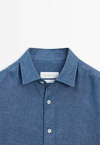 Himmelblaues Leinenhemd mit Kragen und strukturierter Oberfläche, mit einer durchgehenden Knopfleiste und einem Etikett im Kragen. Die Knöpfe sind weiß.