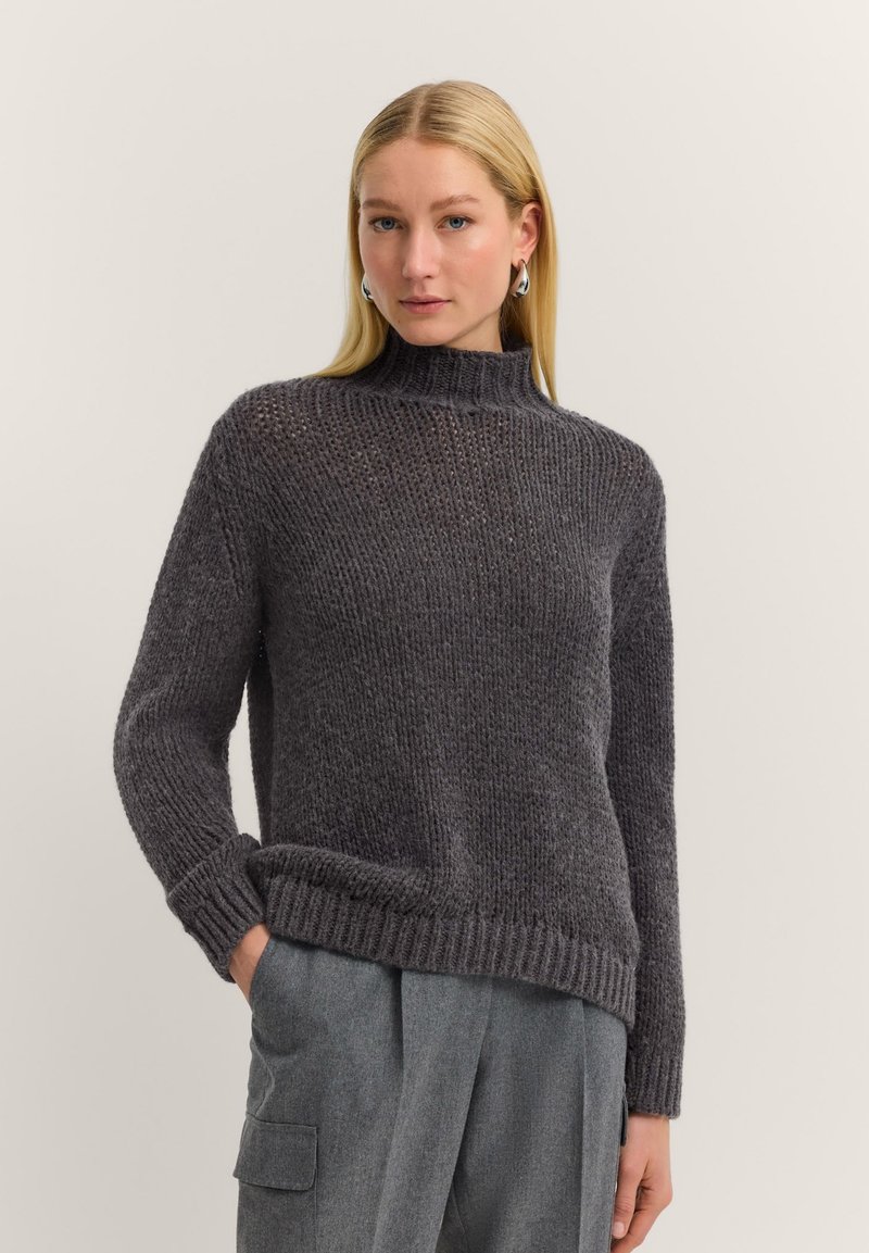 Woman with long blonde hair wears a dark gray chunky knit sweater and gray pants with one hand in pocket against a plain background.