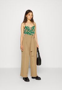 Camisola verde floral com alças finas, combinada com calças largas de cintura alta em bege e sapatos pretos robustos, segurando uma bolsa preta.