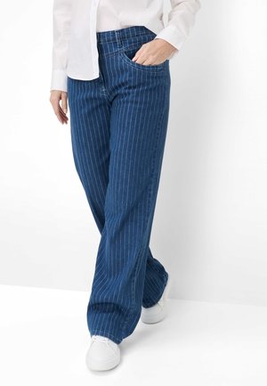 Person iført blå bredbenede bukser med nålestriber, hvid langærmet skjorte og hvide sneakers, med den ene hånd i lommen.