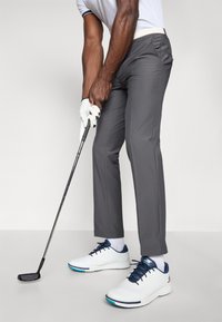 Golf outfit featuring gray pants, a white shirt with navy accents, white shoes with navy and teal details, and a golf club in hand.