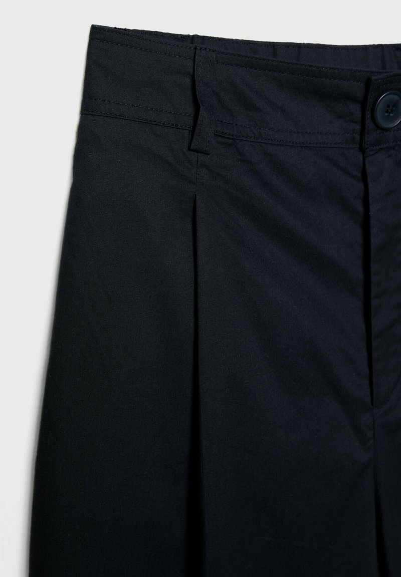 Pantaloni neri in tessuto con pieghe frontali, vita con bottone e texture liscia. Primo piano dei materiali e dei dettagli di design.
