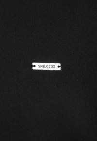 Tessuto nero con una superficie liscia, dotato di una targhetta rettangolare argento incisa "SMILODOX" attaccata alla superficie.