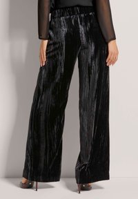 Zwarte wijde broek gemaakt van textuur stof met een glanzende afwerking en verticale plooien. Elastische tailleband en aansluitende boorden bij de enkels.