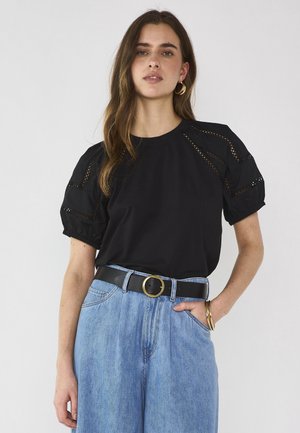 Donna che indossa una maglietta nera a maniche corte con dettagli in pizzo, jeans a vita alta azzurri, cintura nera con fibbia dorata e orecchini a cerchio dorati.