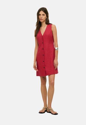 Mujer con vestido rojo sin mangas con botones delanteros, brazalete plateado en el brazo izquierdo, y sandalias planas beige, de pie frente a un fondo liso.