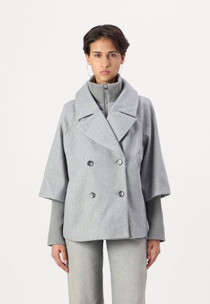 Manteau gris à double boutonnage avec larges revers, manches courtes, tissu texturé et accents de boutons. Porté par-dessus un haut gris à col montant.