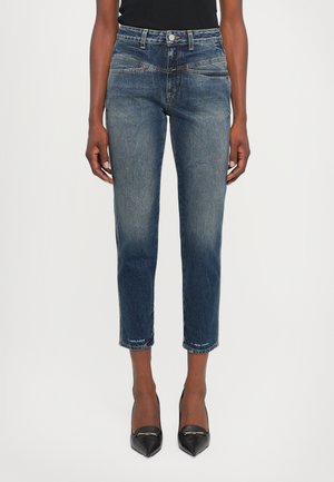 Schmal geschnittene, mittelhohe, blaue verkürzte Jeans, getragen mit schwarzen spitzen Pumps und einem schwarzen Oberteil, gezeigt vom mittleren Oberkörper bis zu den Füßen.